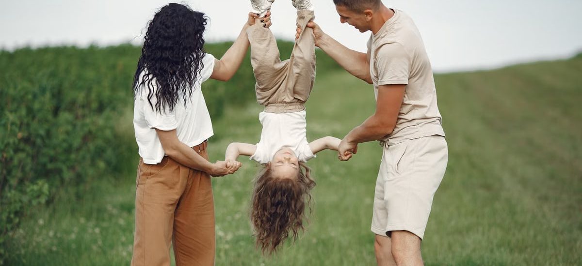 A joyful family enjoys outdoor play, lifting their child upside down in a green field.