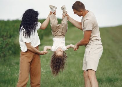 A joyful family enjoys outdoor play, lifting their child upside down in a green field.